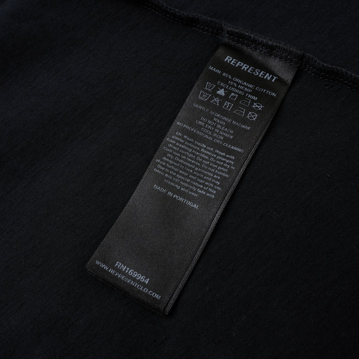 Represent Enemy Tee in washed black, featuring oversized gothic 'Represent' lettering, a hand-drawn lion and diving eagle illustration, blue flame detailing, and 'England' branding on heavyweight cotton