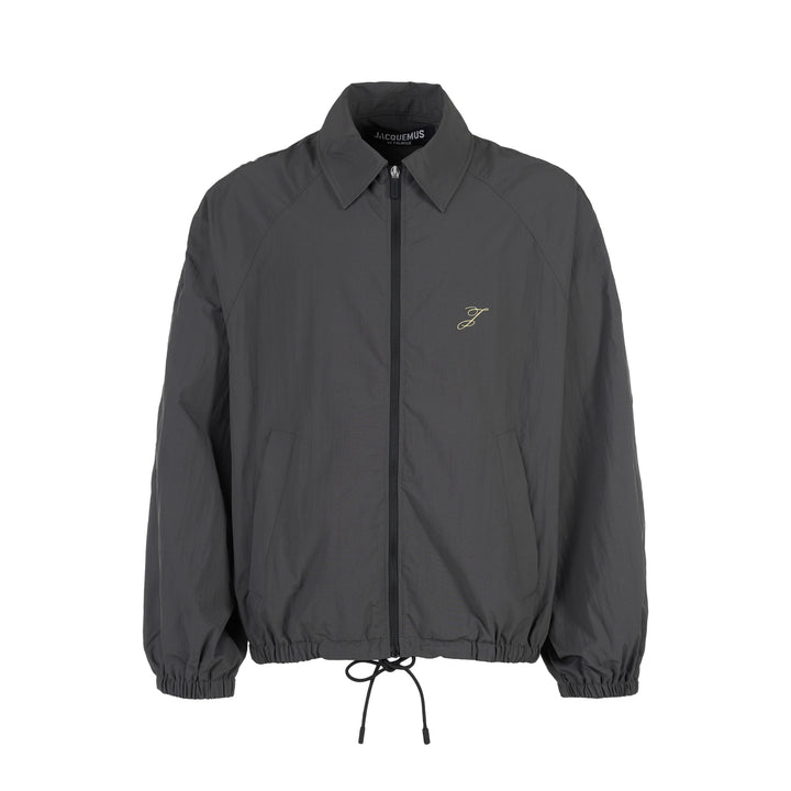 Jacquemus Le Coupe-Vent charcoal grey zip-up windbreaker with collar, elasticated cuffs and hem, and embroidered gold monogram on the chest