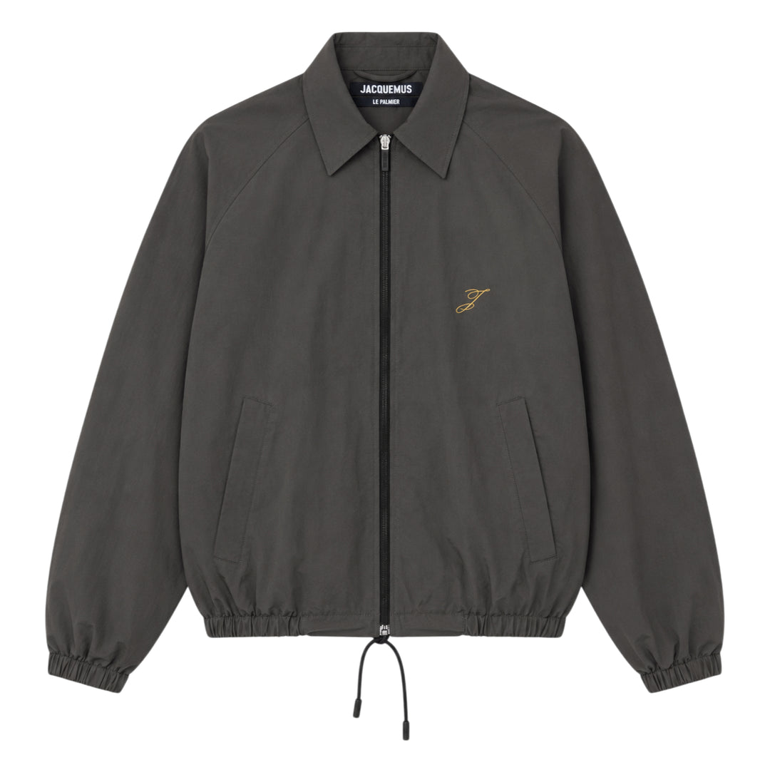 Jacquemus Le Coupe-Vent blouson in slate grey with full-length zip closure, gold script 'J' embroidery on the chest, elasticated cuffs, and drawstring hem