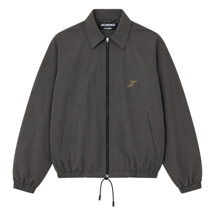Jacquemus Le Coupe-Vent blouson in slate grey with full-length zip closure, gold script 'J' embroidery on the chest, elasticated cuffs, and drawstring hem