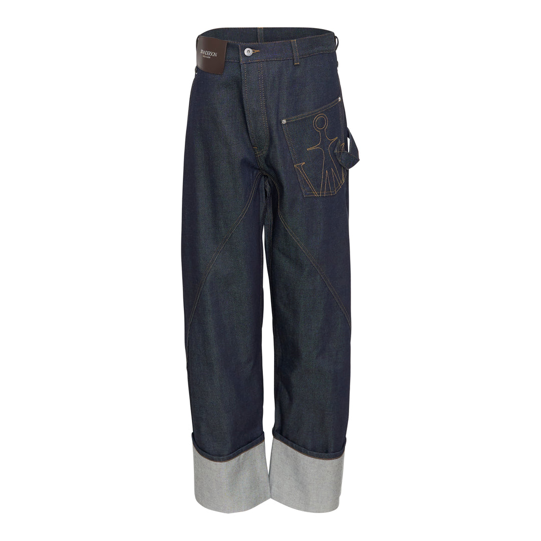 JW Anderson Twisted Workwear Jeans in raw indigo selvedge denim with contrast gold stitching, embroidered figure motif on hip pocket, twisted seam construction, and deep rolled cuffs in grey reverse fabric