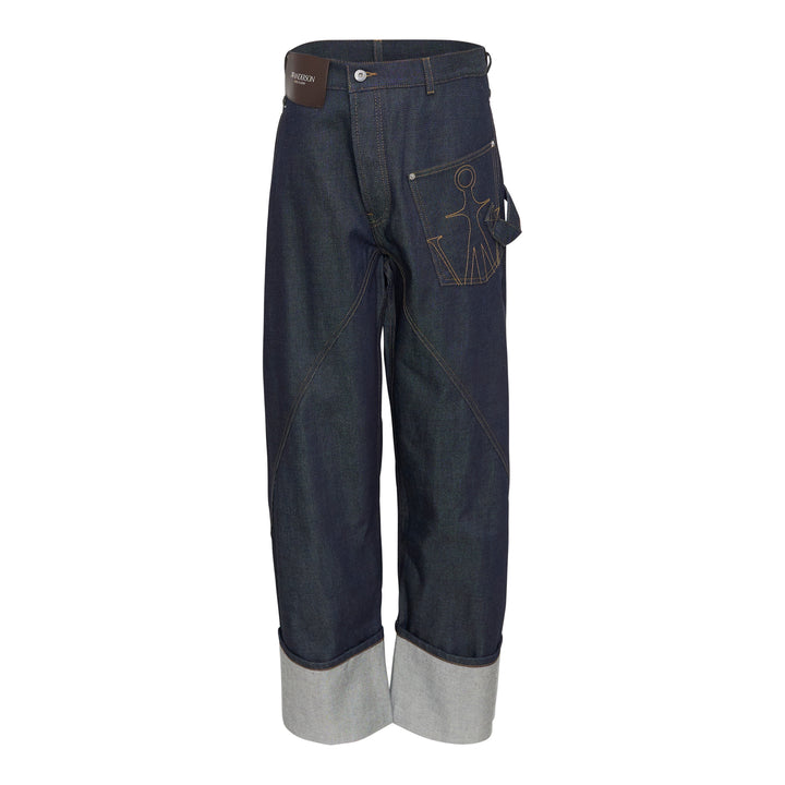 JW Anderson Twisted Workwear Jeans in raw indigo selvedge denim with contrast gold stitching, embroidered figure motif on hip pocket, twisted seam construction, and deep rolled cuffs in grey reverse fabric