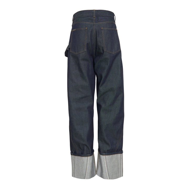 JW Anderson Twisted Workwear Jeans in raw indigo selvedge denim with contrast gold stitching, embroidered figure motif on hip pocket, twisted seam construction, and deep rolled cuffs in grey reverse fabric