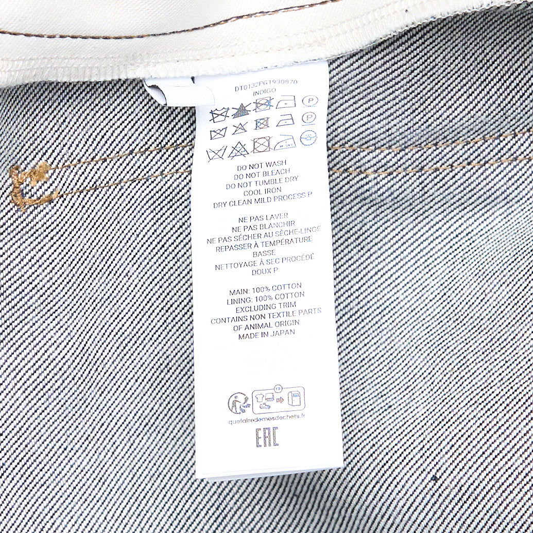 JW Anderson Twisted Workwear Jeans in raw indigo selvedge denim with contrast gold stitching, embroidered figure motif on hip pocket, twisted seam construction, and deep rolled cuffs in grey reverse fabric