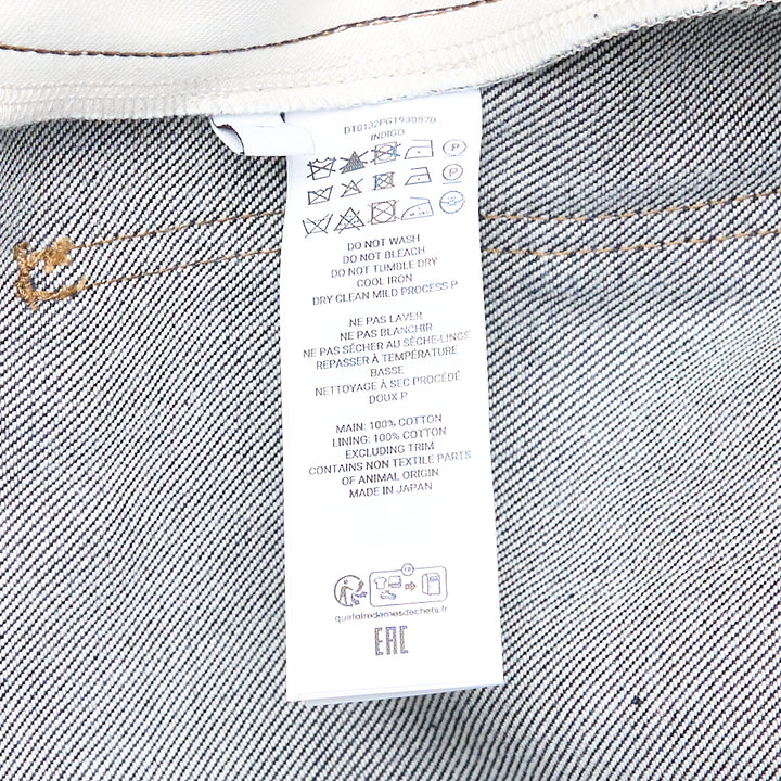 JW Anderson Twisted Workwear Jeans in raw indigo selvedge denim with contrast gold stitching, embroidered figure motif on hip pocket, twisted seam construction, and deep rolled cuffs in grey reverse fabric