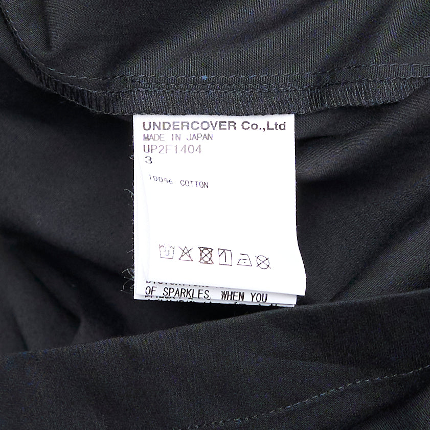 Undercover Cropped U Logo Blouse in black with gathered sleeves, point collar, tonal buttons, and a green embroidered Undercover logo on the chest pocket