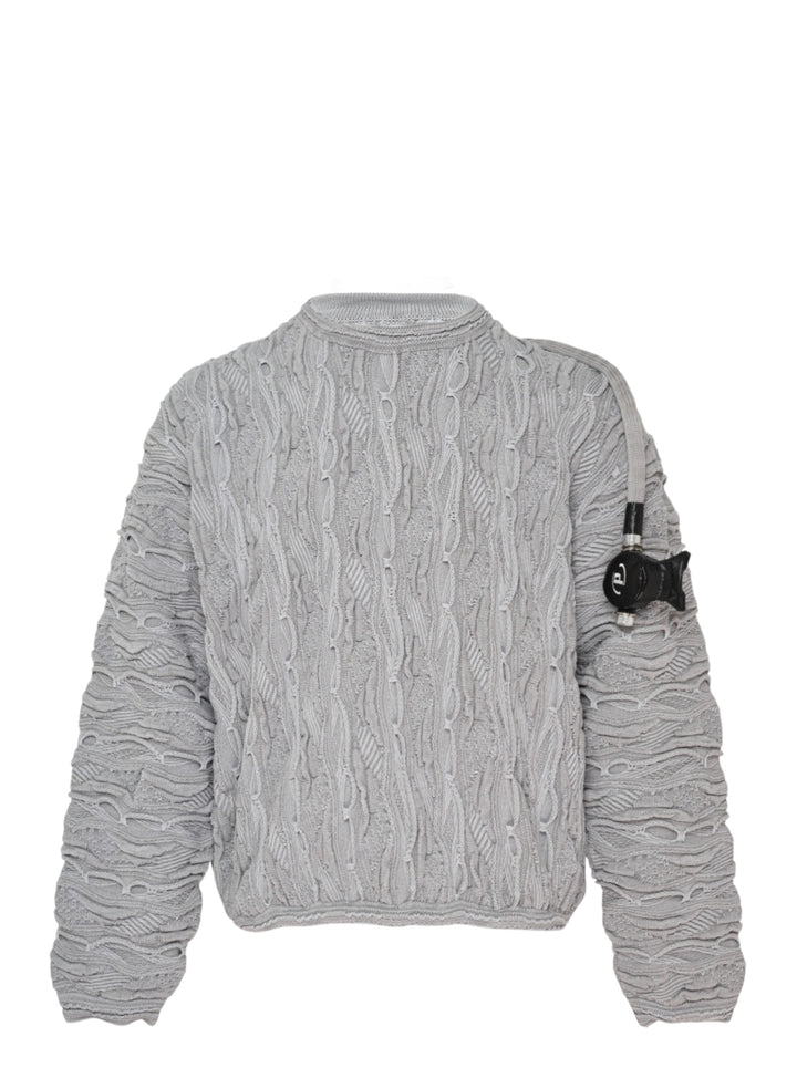 doublet Air Yarn Regulator Knit Pullover in grey, featuring distressed cable-knit texture across the body and sleeves with an attached scuba regulator hardware detail on the sleeve, crew neckline, shot flat on a white background