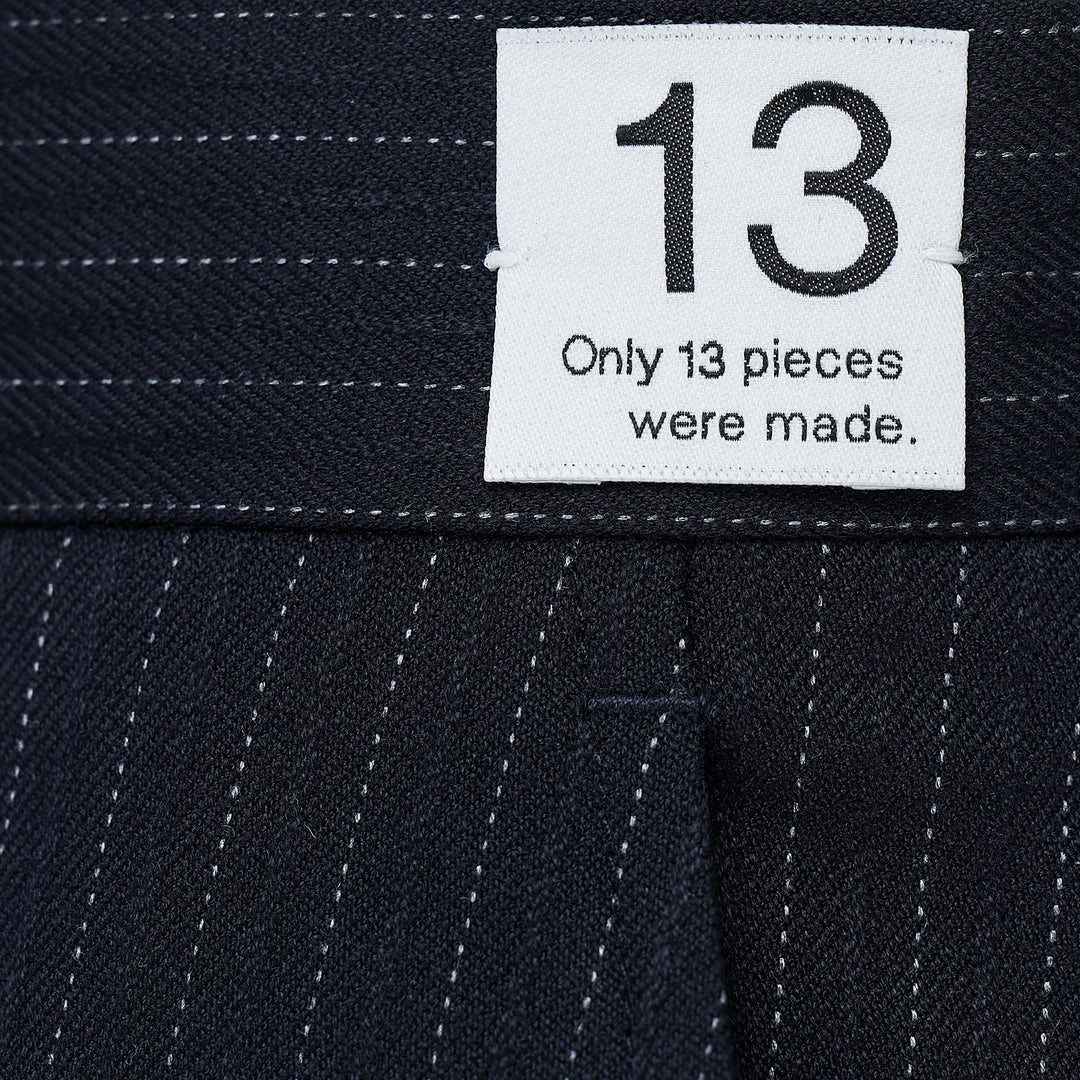 RE;CODE Stripe Side Drape Structured Skirt in navy pinstripe, shown from the side, featuring a tailored silhouette with a draped panel, belt loops, back button pocket, and woven brand label at the waistband