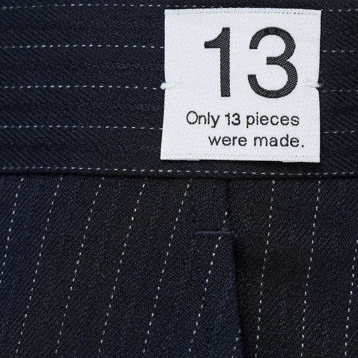RE;CODE Stripe Side Drape Structured Skirt in navy pinstripe, shown from the side, featuring a tailored silhouette with a draped panel, belt loops, back button pocket, and woven brand label at the waistband