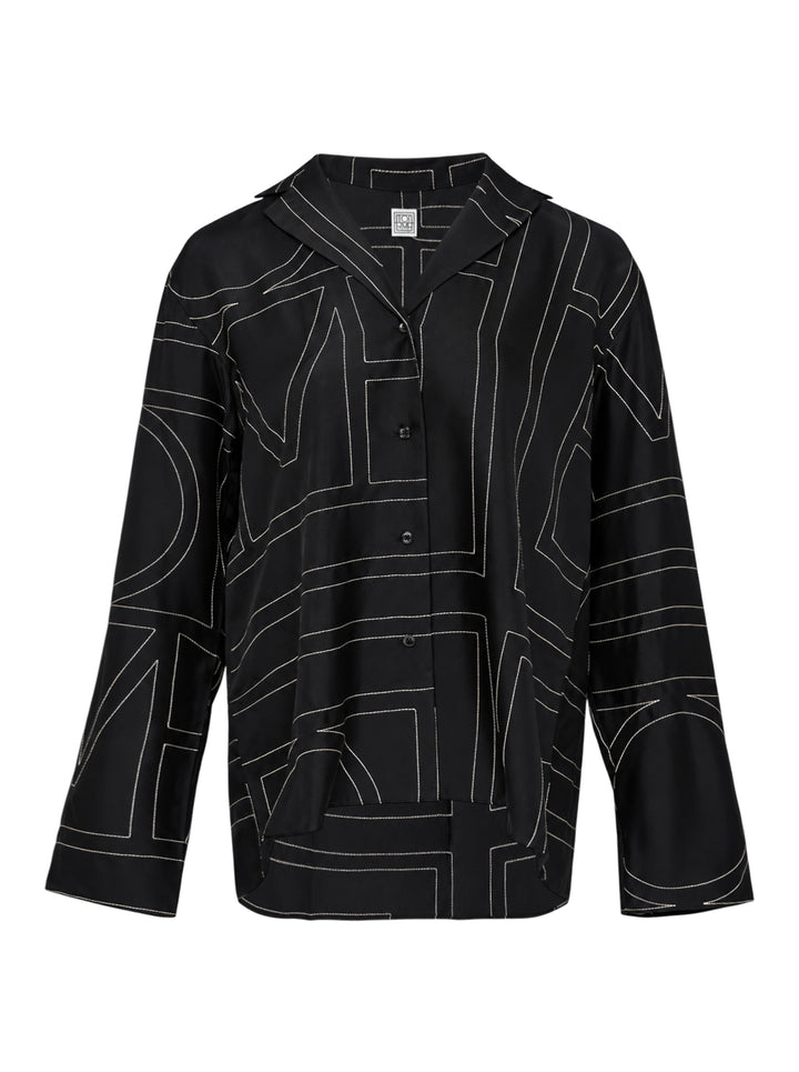 Totême Monogram Silk Pyjama Top in black, featuring all-over contrast-stitched monogram print, camp collar, partial button placket, and relaxed long-sleeve silhouette