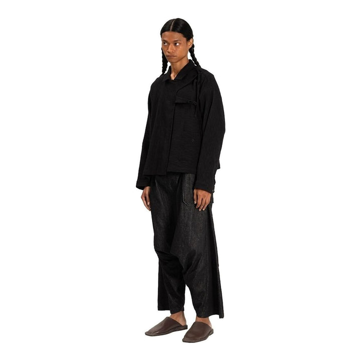 Model wearing Anaabu Asymmetrical Batwing Top in black textured fabric with an open collar, asymmetric front placket, patch chest pocket, and long batwing sleeves, styled with dark drop-crotch trousers and brown mules - packshot