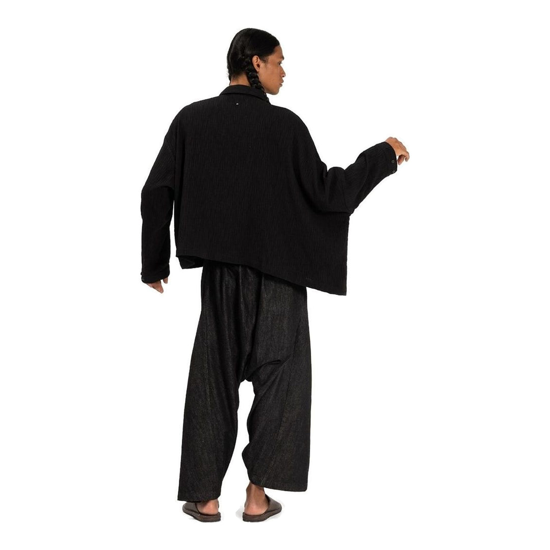 Model wearing Anaabu Asymmetrical Batwing Top in black textured fabric with an open collar, asymmetric front placket, patch chest pocket, and long batwing sleeves, styled with dark drop-crotch trousers and brown mules - packshot