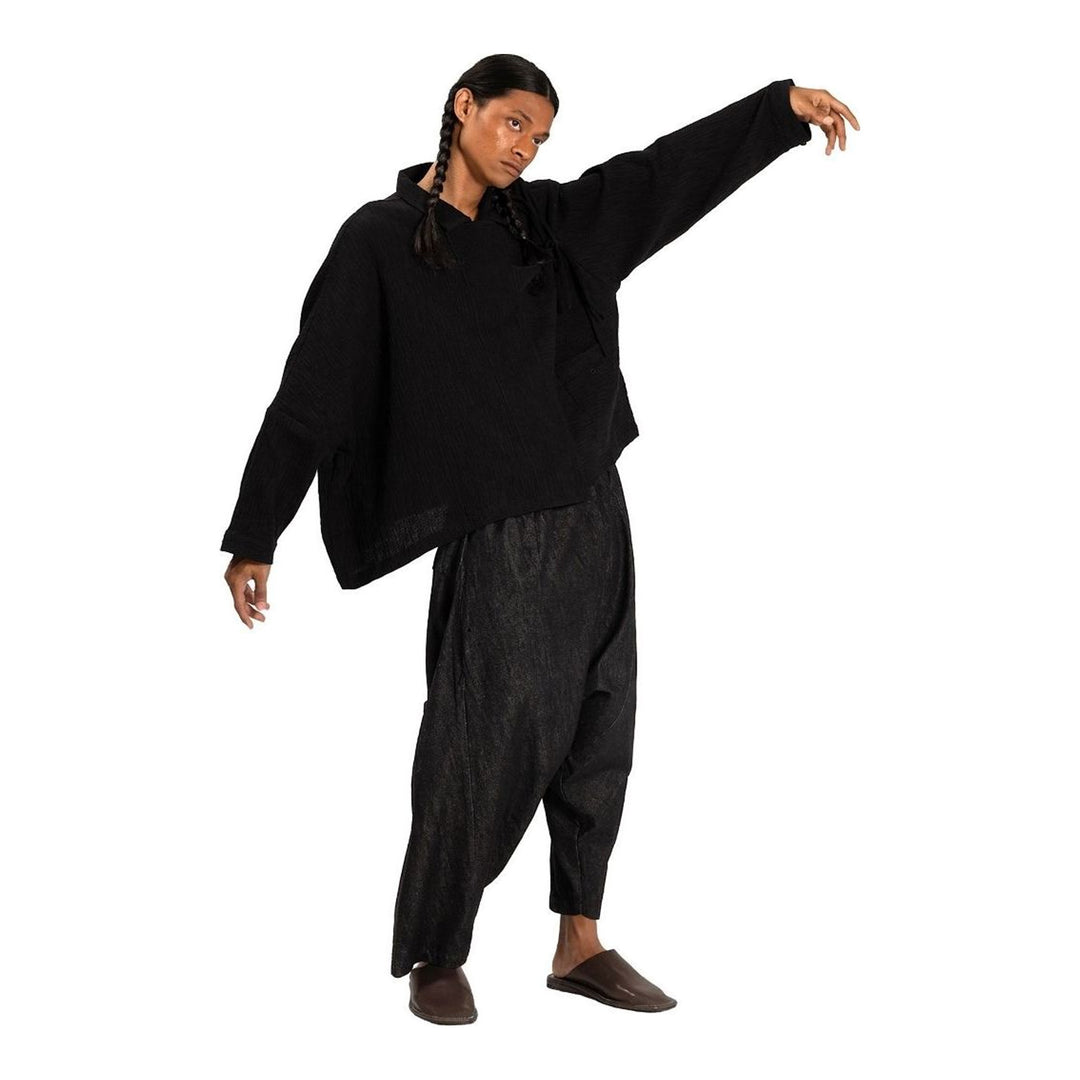 Model wearing Anaabu Asymmetrical Batwing Top in black textured fabric with an open collar, asymmetric front placket, patch chest pocket, and long batwing sleeves, styled with dark drop-crotch trousers and brown mules - packshot