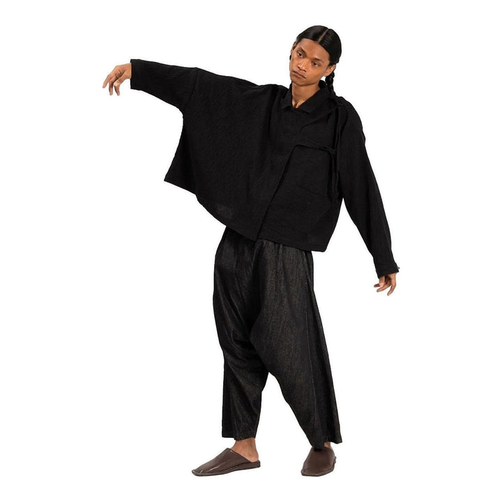 Model wearing Anaabu Asymmetrical Batwing Top in black textured fabric with an open collar, asymmetric front placket, patch chest pocket, and long batwing sleeves, styled with dark drop-crotch trousers and brown mules - packshot