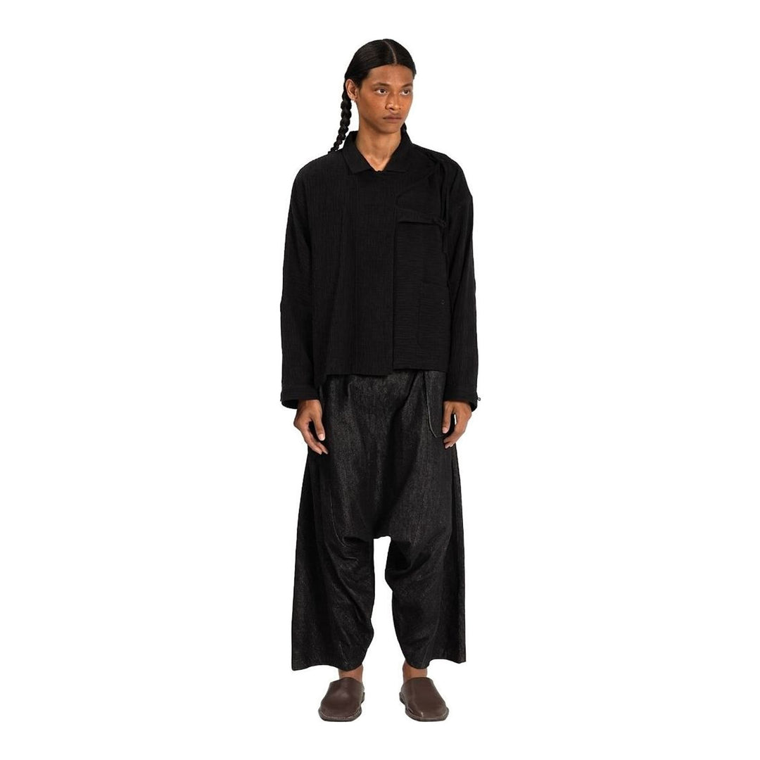 Model wearing Anaabu Asymmetrical Batwing Top in black textured fabric with an open collar, asymmetric front placket, patch chest pocket, and long batwing sleeves, styled with dark drop-crotch trousers and brown mules - packshot