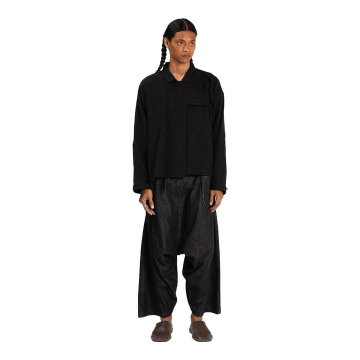 Model wearing Anaabu Asymmetrical Batwing Top in black textured fabric with an open collar, asymmetric front placket, patch chest pocket, and long batwing sleeves, styled with dark drop-crotch trousers and brown mules - packshot