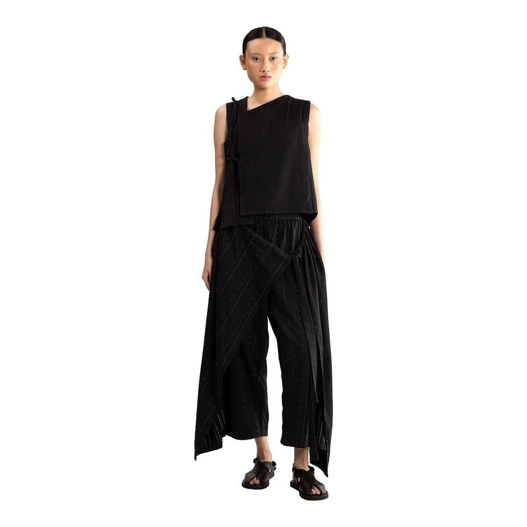 Model wearing the Anaabu Asymmetrical Vest in black, featuring an asymmetric neckline, diagonal panelling, and a draped fringe tie at the shoulder, styled with wide-leg black trousers and dark leather sandals. - packshot