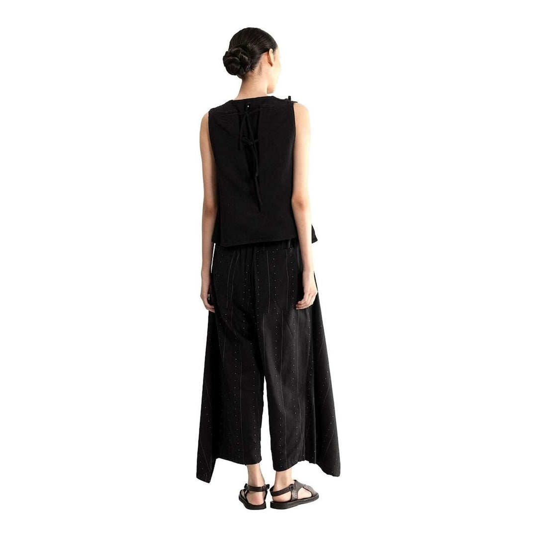 Model wearing the Anaabu Asymmetrical Vest in black, featuring an asymmetric neckline, diagonal panelling, and a draped fringe tie at the shoulder, styled with wide-leg black trousers and dark leather sandals. - packshot
