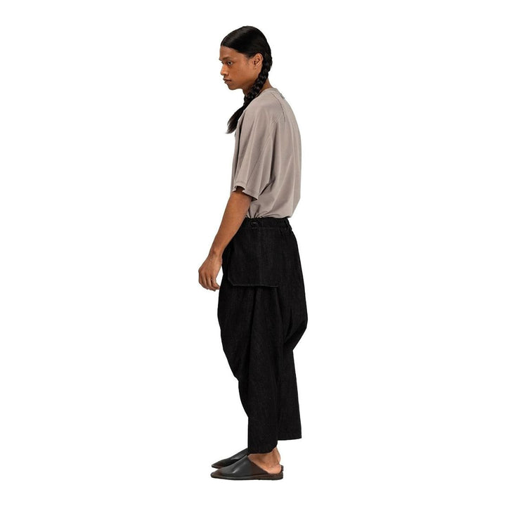 Model wearing Anaabu Dropped Culotte Pants in black — wide-leg drop-crotch trousers with an asymmetric draped skirt overlay panel, styled with a black crop top, frayed bucket hat, and tabi sneakers - packshot