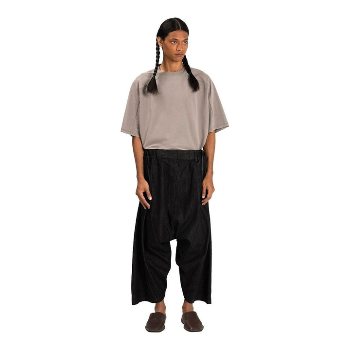Model wearing Anaabu Dropped Culotte Pants in black — wide-leg drop-crotch trousers with an asymmetric draped skirt overlay panel, styled with a black crop top, frayed bucket hat, and tabi sneakers - packshot