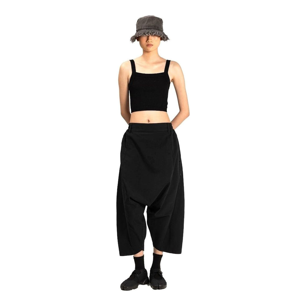 Model wearing Anaabu Dropped Culotte Pants in black — wide-leg drop-crotch trousers with an asymmetric draped skirt overlay panel, styled with a black crop top, frayed bucket hat, and tabi sneakers - packshot