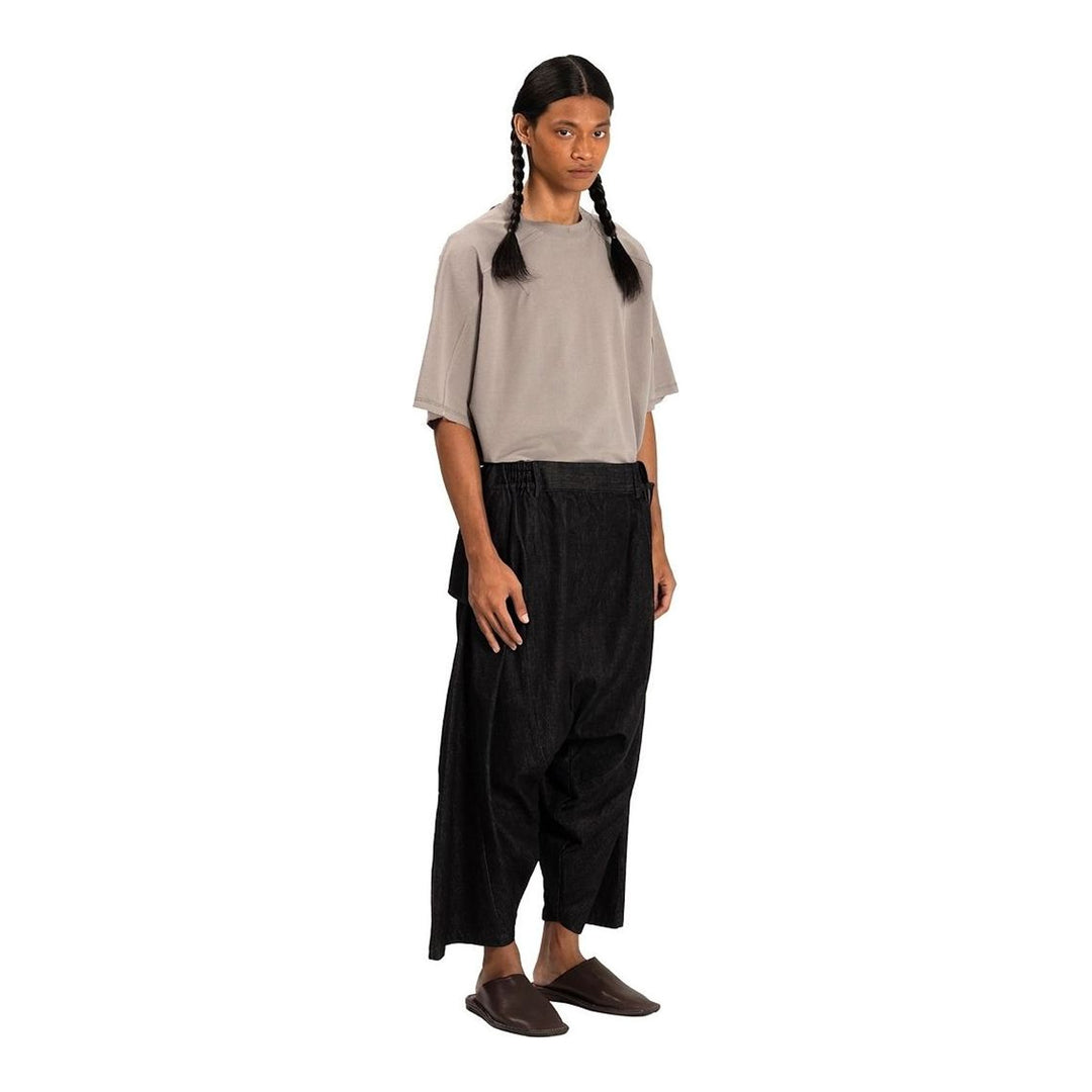 Model wearing Anaabu Dropped Culotte Pants in black — wide-leg drop-crotch trousers with an asymmetric draped skirt overlay panel, styled with a black crop top, frayed bucket hat, and tabi sneakers - packshot
