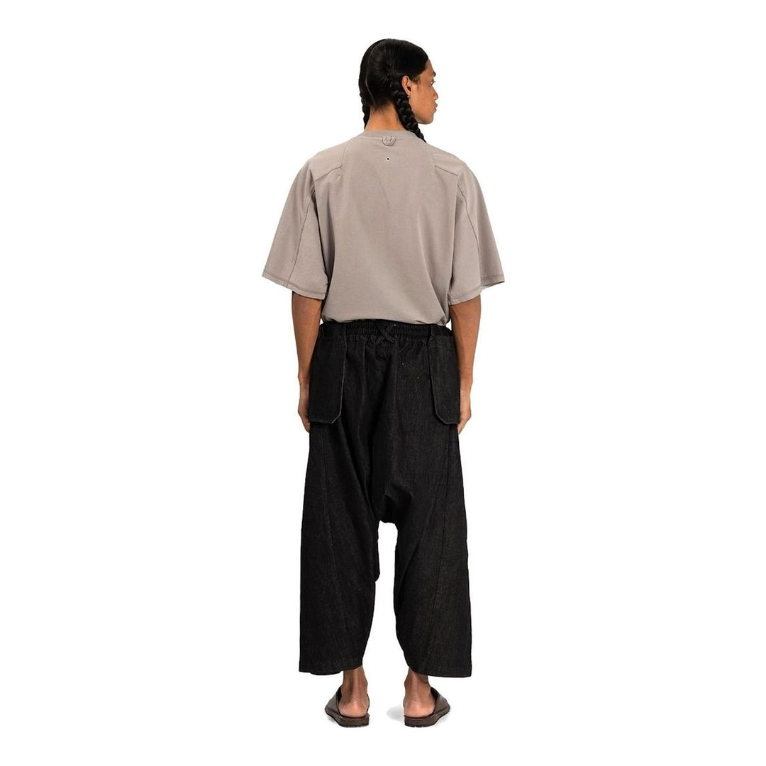 Model wearing Anaabu Dropped Culotte Pants in black — wide-leg drop-crotch trousers with an asymmetric draped skirt overlay panel, styled with a black crop top, frayed bucket hat, and tabi sneakers - packshot