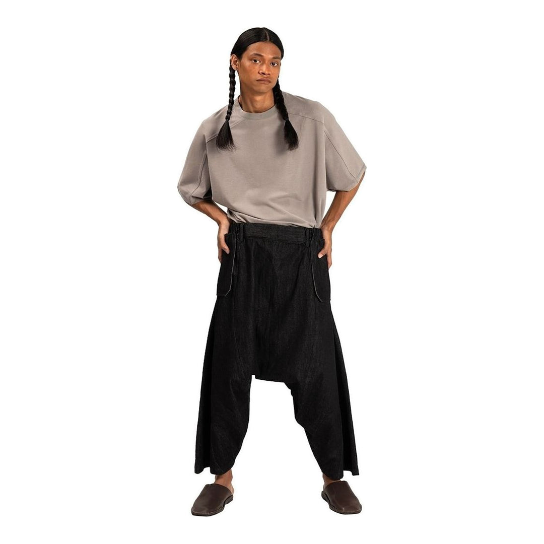 Model wearing Anaabu Dropped Culotte Pants in black — wide-leg drop-crotch trousers with an asymmetric draped skirt overlay panel, styled with a black crop top, frayed bucket hat, and tabi sneakers - packshot