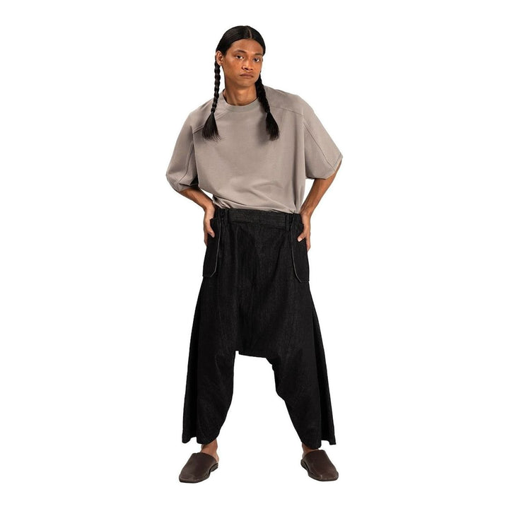Model wearing Anaabu Dropped Culotte Pants in black — wide-leg drop-crotch trousers with an asymmetric draped skirt overlay panel, styled with a black crop top, frayed bucket hat, and tabi sneakers - packshot