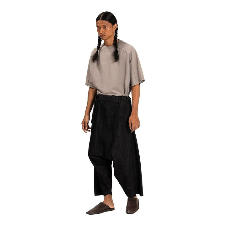 Model wearing Anaabu Dropped Culotte Pants in black — wide-leg drop-crotch trousers with an asymmetric draped skirt overlay panel, styled with a black crop top, frayed bucket hat, and tabi sneakers - packshot