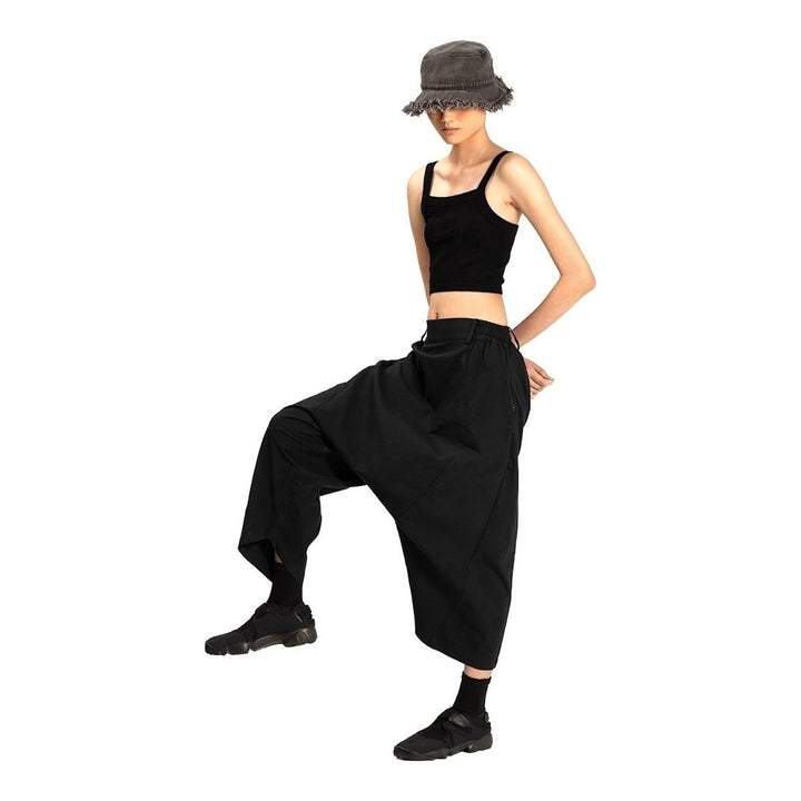 Model wearing Anaabu Dropped Culotte Pants in black — wide-leg drop-crotch trousers with an asymmetric draped skirt overlay panel, styled with a black crop top, frayed bucket hat, and tabi sneakers - packshot