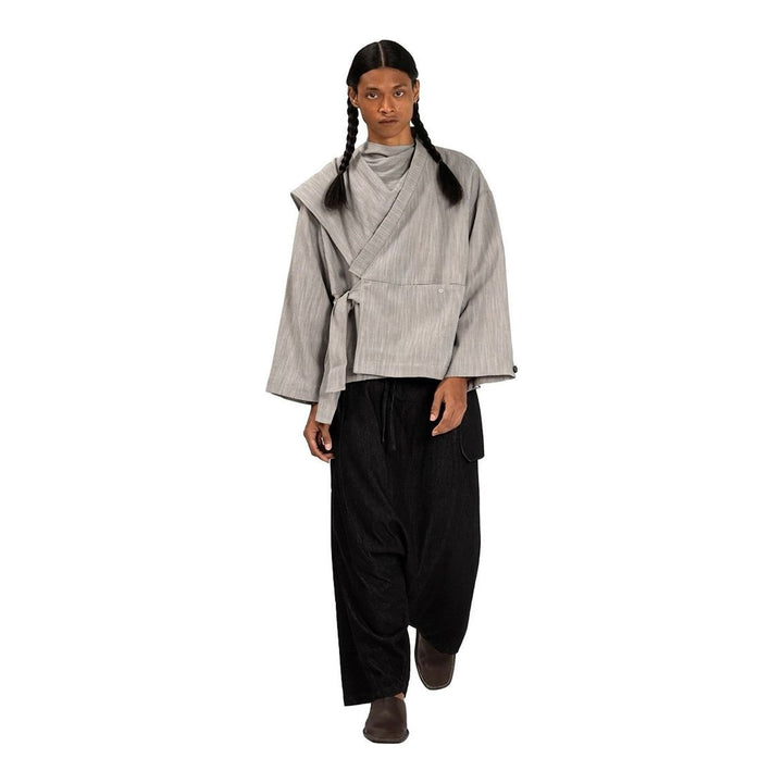 Model wearing the Anaabu Shawl Jacket in black — a draped wrap-style jacket with oversized shawl collar and self-tie belt, styled over a black turtleneck and olive wide-leg trousers - packshot