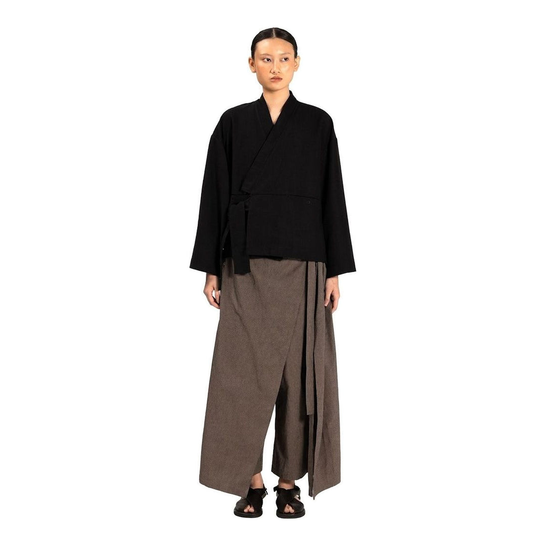 Model wearing the Anaabu Shawl Jacket in black — a draped wrap-style jacket with oversized shawl collar and self-tie belt, styled over a black turtleneck and olive wide-leg trousers - packshot