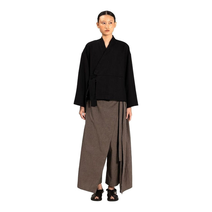 Model wearing the Anaabu Shawl Jacket in black — a draped wrap-style jacket with oversized shawl collar and self-tie belt, styled over a black turtleneck and olive wide-leg trousers - packshot